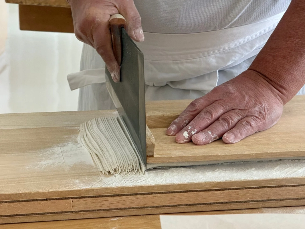 職人が木製の台の上で、大きな包丁を使って手打ちの麺を丁寧に切っている様子