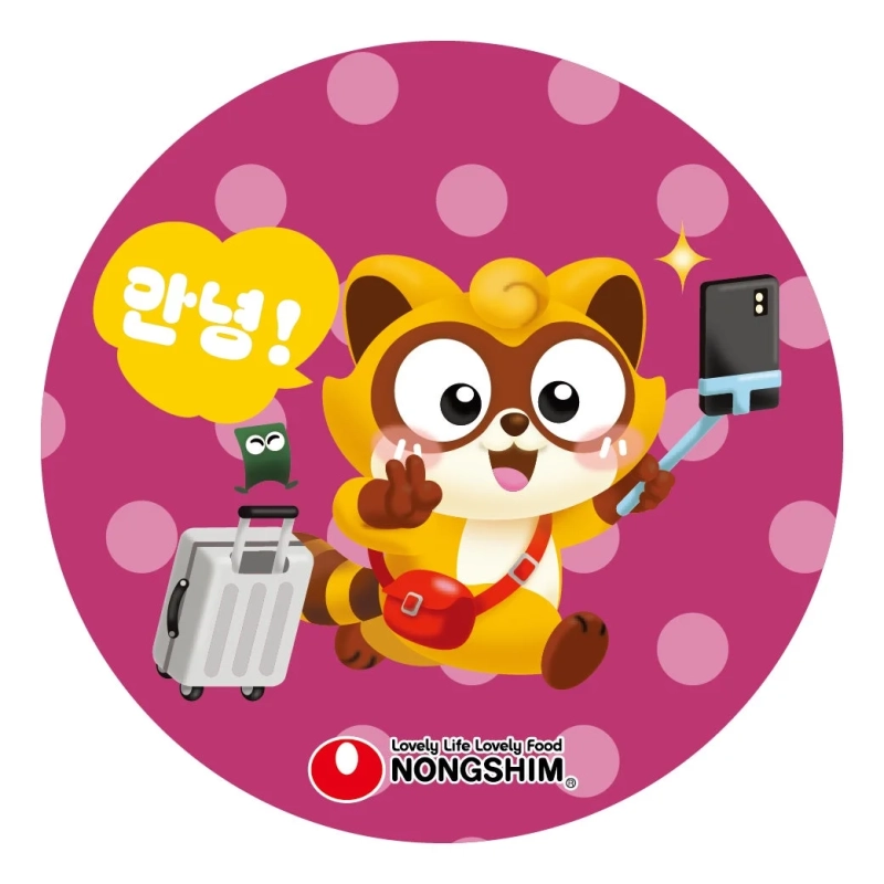 ピンクの水玉模様の背景に、農心のタヌキのようなキャラクターがピースサインをして自撮りをするイラストです。旅行鞄と小さな緑の生き物も描かれ、「アンニョン！」と挨拶する楽しげな旅行気分を表現しています。