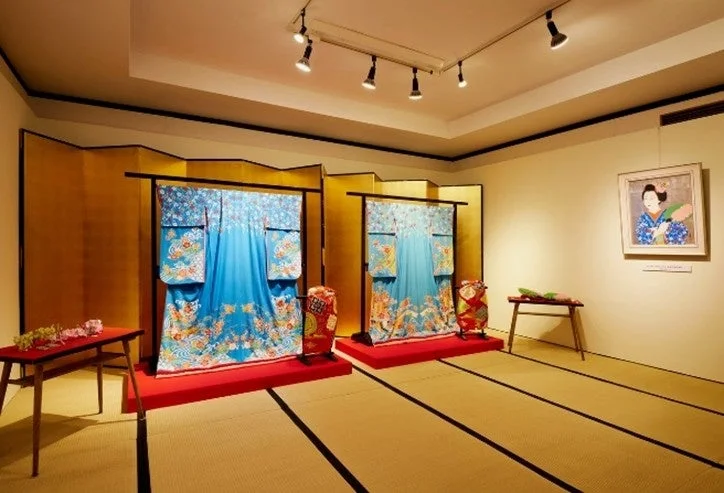 伝統的な日本文化の展示空間で、豪華な青い着物二点が金屏風を背景に展示されています。畳敷きの室内には、壁に絵画が飾られ、装飾品が置かれたテーブルも見えます。