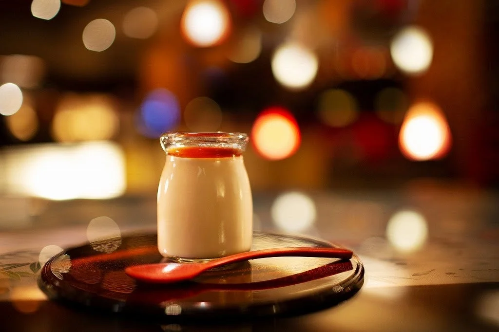 温かいボケの光を背景に、ガラス瓶に入ったプリンかパンナコッタが赤いスプーンと共に黒いトレイに置かれている