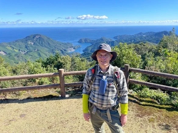 晴れた日に山頂らしき場所で景色を背景に立つ男性の姿