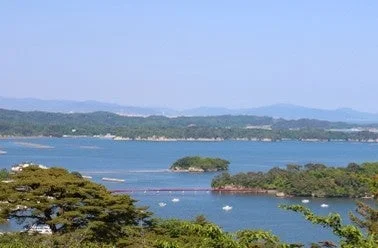 複数の島々が浮かぶ穏やかな湾の風景。中央には小さな島々を結ぶ赤い橋が見え、水面には白い船が点在しています。