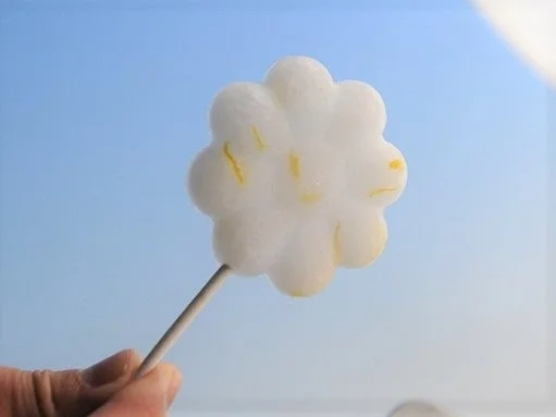 青い空を背景に、白い花のような形をしたふわふわのお菓子が棒に刺さっており、それを手が持っています