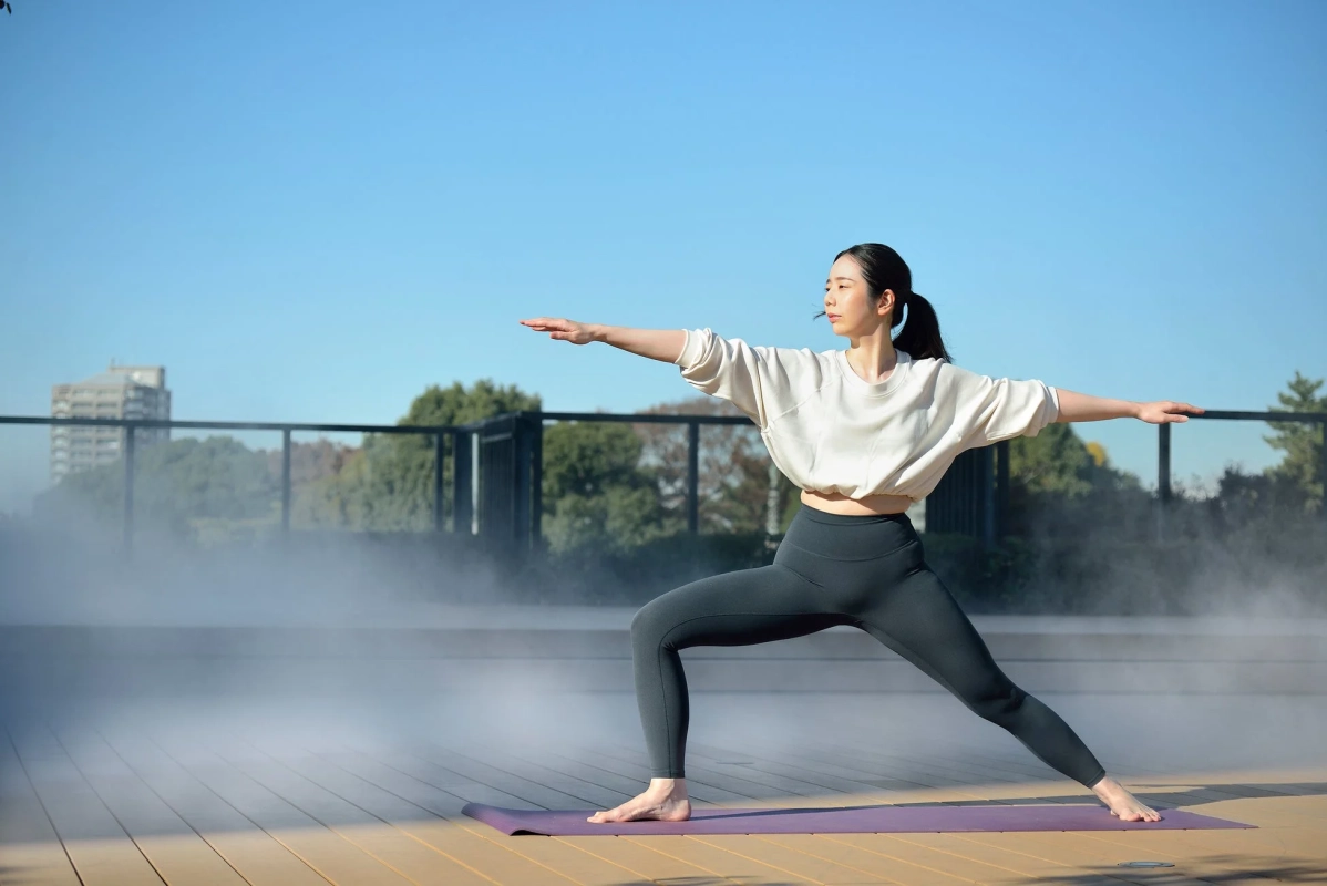 青空の下、屋外のウッドデッキでヨガのポーズ（ウォーリアII）をとる女性の姿です。足元には霧が立ち込め、穏やかな雰囲気の中で運動しています。