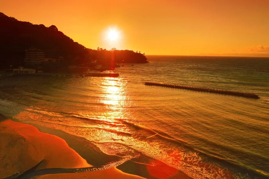 黄金色の夕日が海面に反射し、波打ち際を照らす美しい光景です。左手には山と建物が見え、沖には防波堤が伸びています。穏やかな夕暮れのビーチの景色が広がっています。