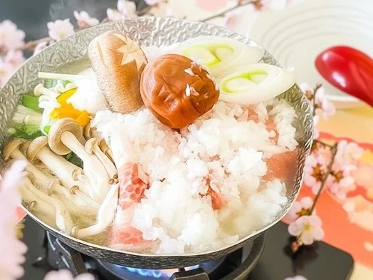 桜の装飾が施された食卓で、肉、きのこ、長ねぎ、大根おろし、梅干しなど具材たっぷりの鍋料理が煮込まれている