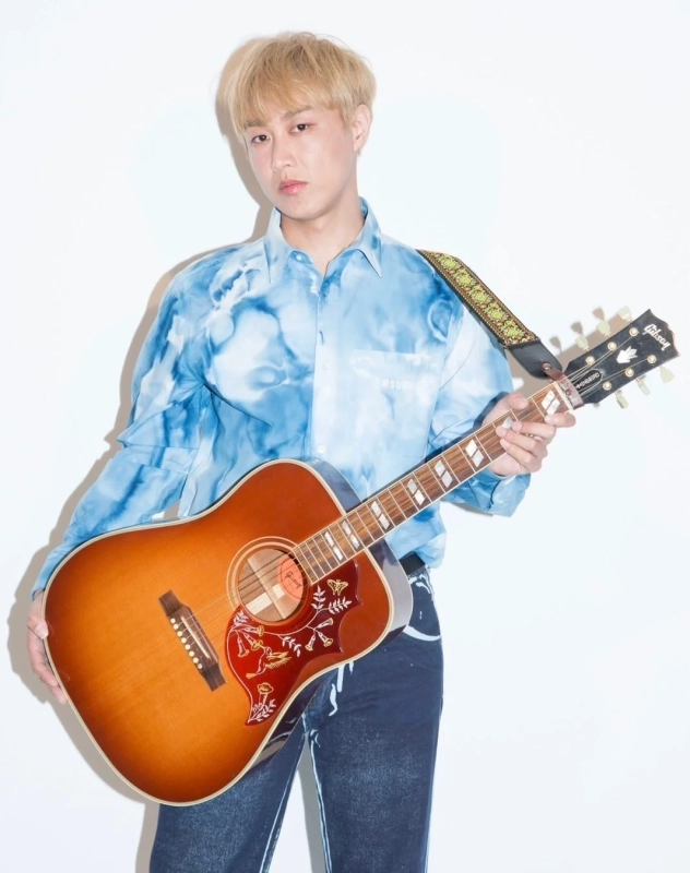 青いタイダイ柄のシャツを着てアコースティックギターを抱える金髪の若い男性
