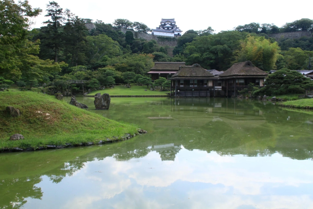 緑豊かな日本庭園の池を中心に、水面に伝統的な建物と遠くの丘に立つ城が映り込む美しい風景です。静寂な雰囲気が漂い、歴史と自然が調和した日本の美を感じさせます。
