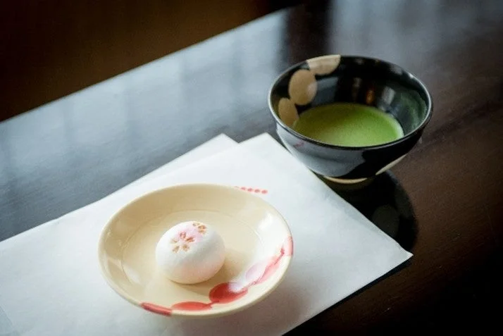 黒い茶碗に入った鮮やかな緑色の抹茶と、桜の模様が描かれた白い和菓子が皿に置かれています。日本の伝統的なお茶の時間を思わせる、静かで美しい風景です。