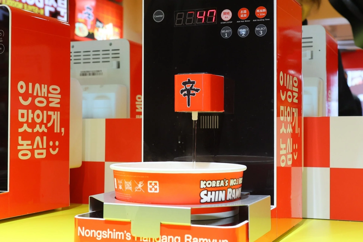 辛ラーメンのカップ麺がセットされた自動調理器で、お湯が注がれている様子を捉えた画像です。韓国語や日本語の操作パネルがあり、手軽にラーメンが楽しめるサービスが提供されていることがわかります。