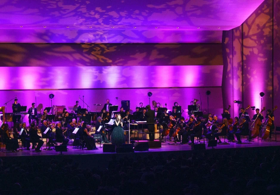 紫色の照明が印象的なステージで、女性歌手がオーケストラをバックに歌唱しているライブパフォーマンスの様子