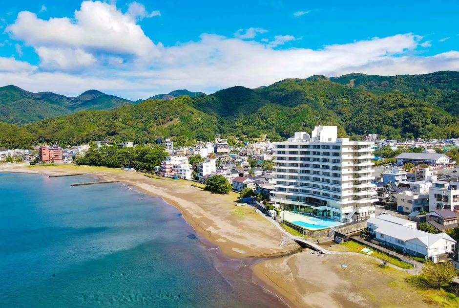 青い海と砂浜が広がる海岸線に白い大型ホテルが建ち並び、背後には緑豊かな山々が連なる美しい風景。晴れた空には白い雲が浮かび、リゾート地のような穏やかな雰囲気が漂う航空写真です。