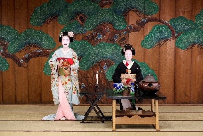 伝統的な着物を着用し、白い化粧をした二人の女性が写っています。一人は立ち、もう一人は茶道具のあるテーブルの前に座っています。松の絵が描かれた背景の前で、日本の伝統文化を表現しているようです。