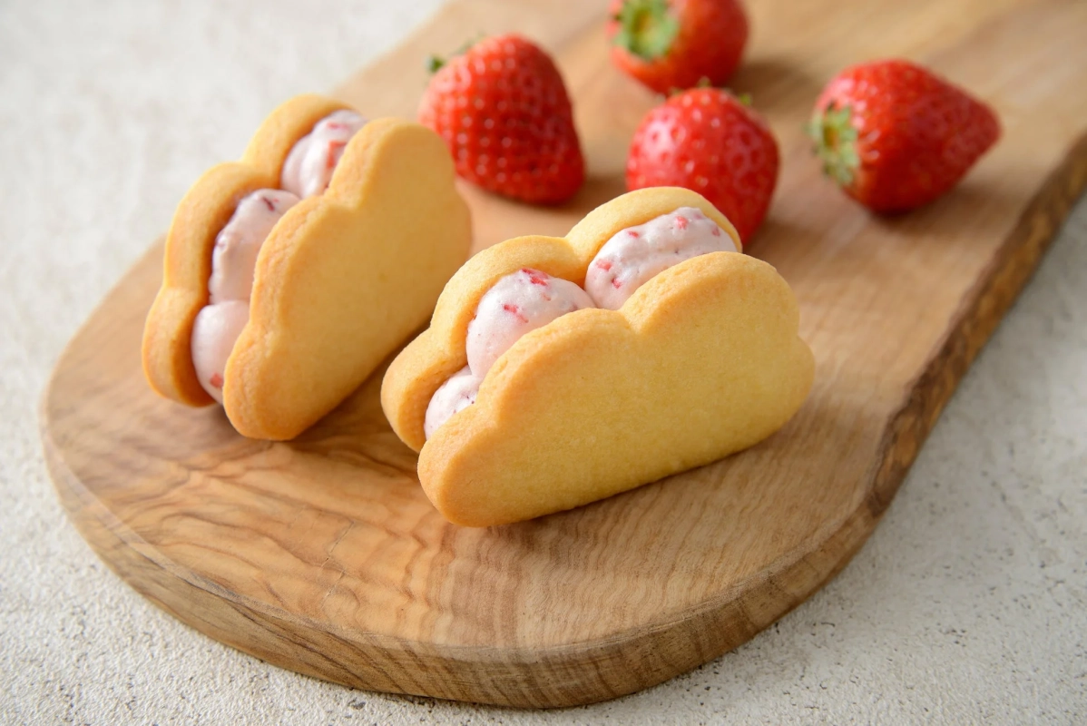 木製のボードの上に、雲の形をしたクッキーサンドが2つ並べられています。クッキーの間にはピンク色のクリームが挟まっており、背景には数個の新鮮ないちごが散りばめられています。