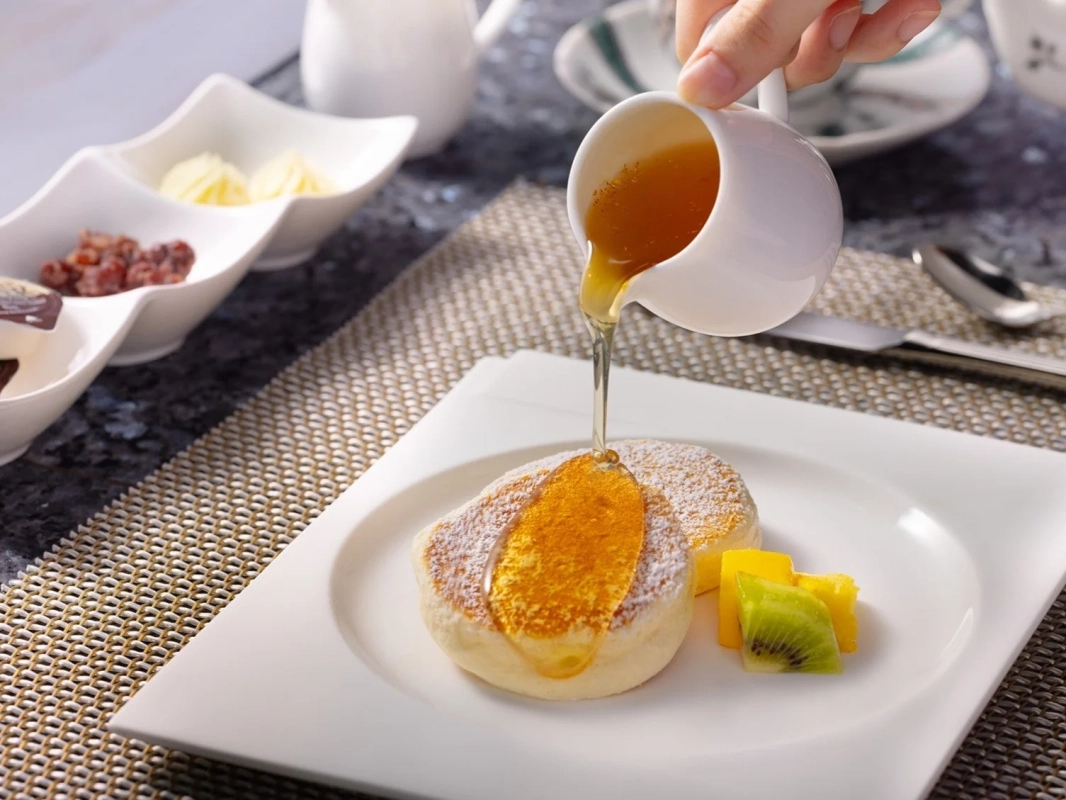 手でシロップをかけているふわふわのスフレパンケーキがメインの画像