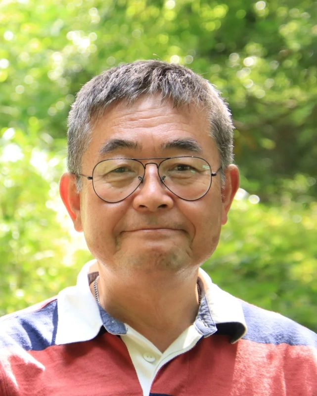 眼鏡をかけたアジア系の高齢男性が、緑豊かな屋外を背景に、カジュアルなポロシャツを着て微笑んでいるポートレート写真です。穏やかな表情が印象的です。