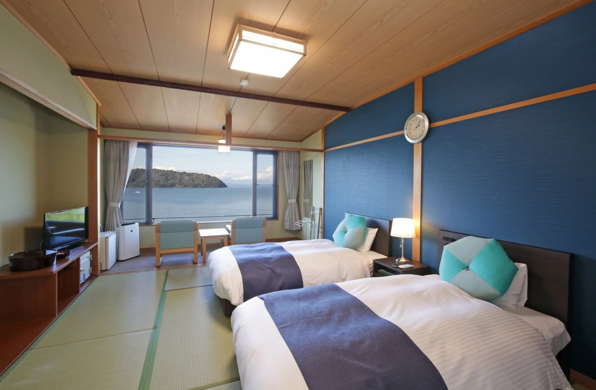 海が見える広々とした和洋室の寝室です。ツインベッドが2台置かれ、窓からは美しい海と島を望むことができます。室内は畳敷きのエリアもあり、青い壁と木目の天井が特徴的で、モダンで落ち着いた雰囲気です。