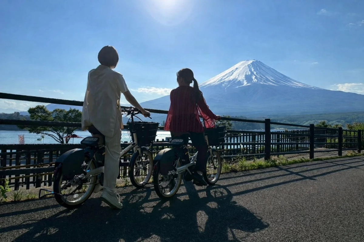 晴れた日に湖畔で自転車に乗る二人が、雪化粧した雄大な富士山を眺めている美しい風景写真です。