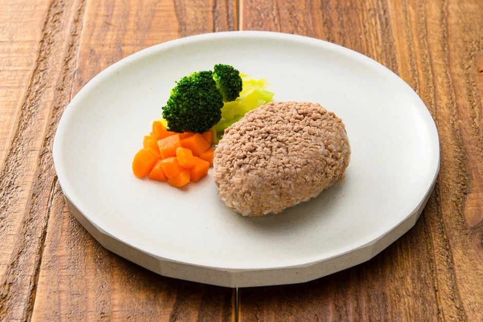 白い皿に盛り付けられたハンバーグと、付け合わせのブロッコリー、人参、そして淡い緑色の野菜が写っています。木目のテーブルに置かれた健康的な食事の風景です。