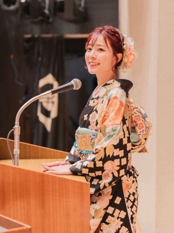 華やかな着物を着た女性が演台の前に立ち、マイクに向かって笑顔で話している