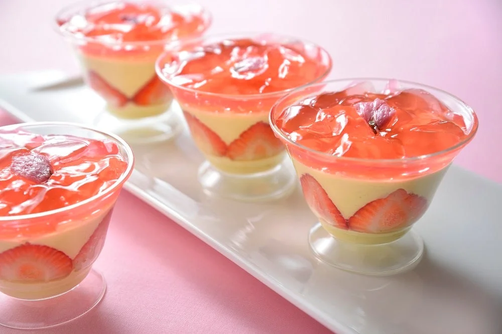 白いプレートに並べられた複数のカップデザート。スライスされたイチゴ、クリーミーな層、そしてピンク色のゼリーで構成されており、ゼリーの上には桜の花が添えられています。