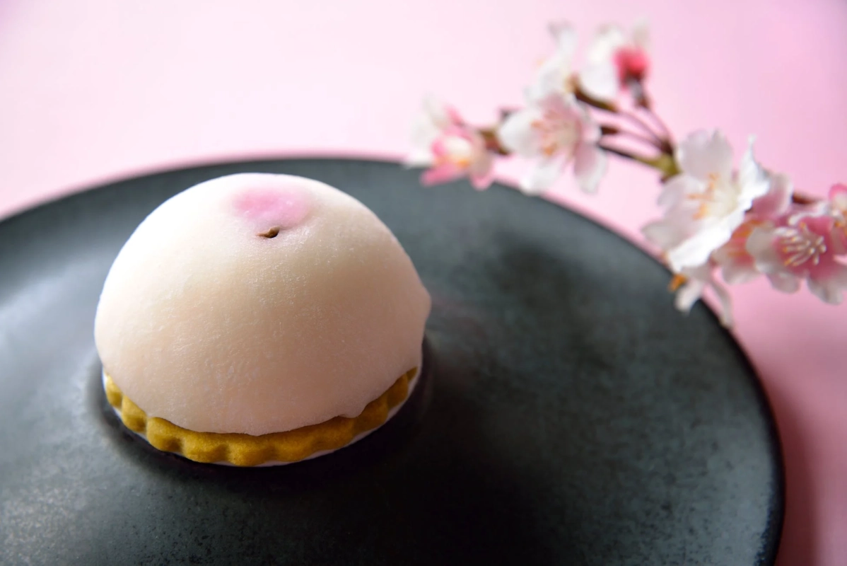暗い皿の上に置かれた、桃のような形をした白い和菓子。頂点には淡いピンク色の模様があり、背景にはぼんやりと桜の花が写り、春の訪れを感じさせる一枚です。