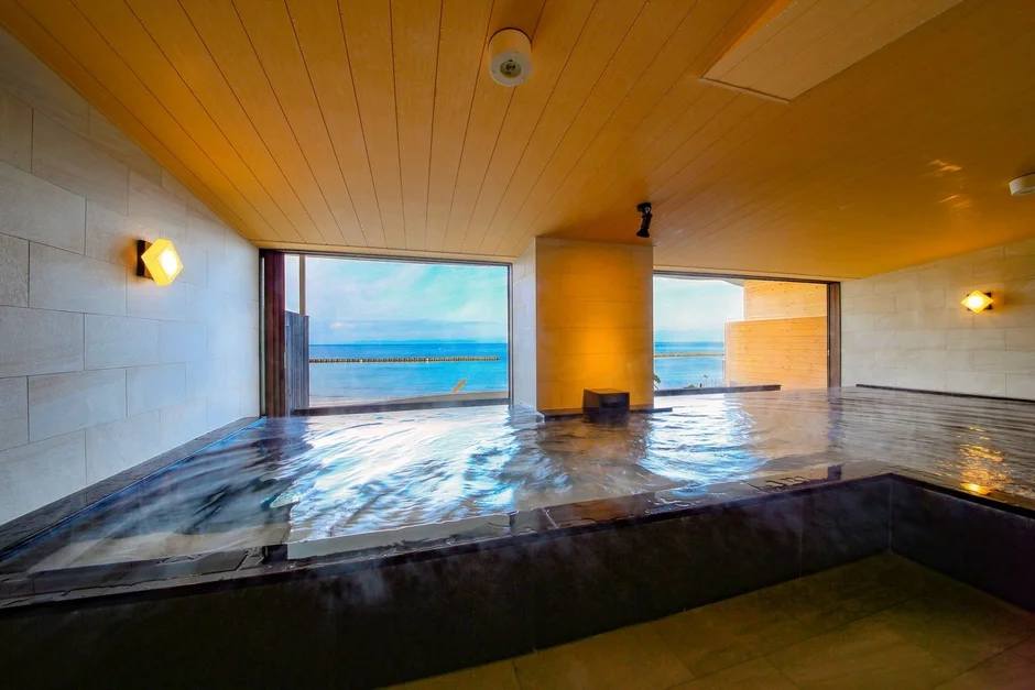 海に面した広々とした屋内風呂で、湯気が立ち上る浴槽が特徴的です。大きな窓からは青い海と空が広がり、リラックスできる開放的な空間が演出されています。