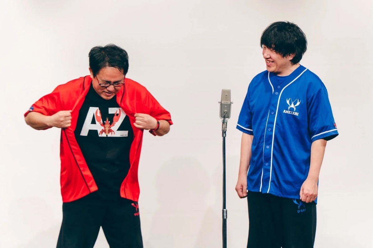 白い背景のスタジオで、男性が二人並んで立っています。一人は赤いシャツの下にロブスター柄のTシャツを見せ、もう一人は青い野球シャツを着ています。中央にはマイクが置かれており、二人は楽しそうに笑っている様子