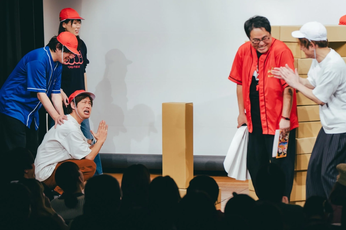 舞台上で数人がパフォーマンスを行っており、赤い帽子をかぶった男性が大げさな表情でしゃがみこんでいます。