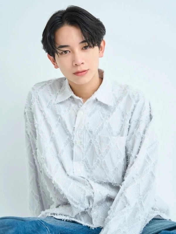 白い背景の前で、白い独特なデザインのシャツとジーンズを着用した若いアジア人男性がカメラを見つめているポートレート写真です。