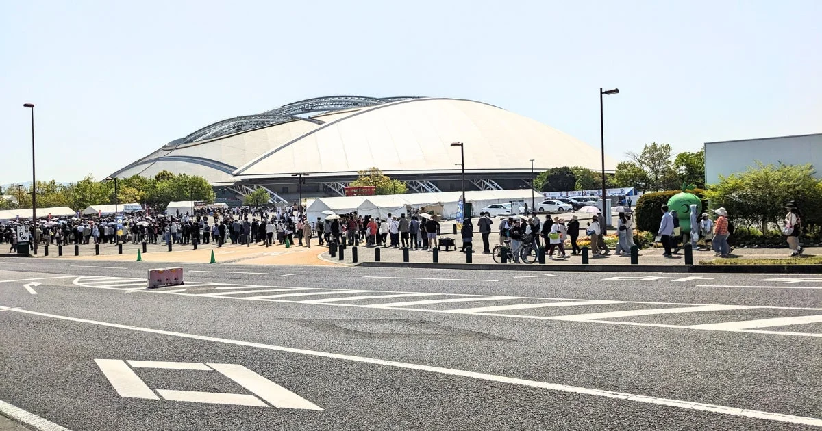 晴れた日に、多くの人々がドーム型スタジアムの前に集まり、イベントの開始を待っている様子。白いテントや緑色のマスコットも見られる。