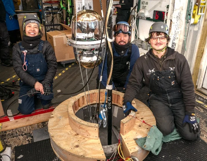 船上または研究施設内で、3人の研究者らしき人々が笑顔でカメラにポーズをとっている。彼らは大きな金色の球体型装置を囲んでおり、その装置は床の開口部から吊り下げられている。研究活動の様子を捉えた一枚。