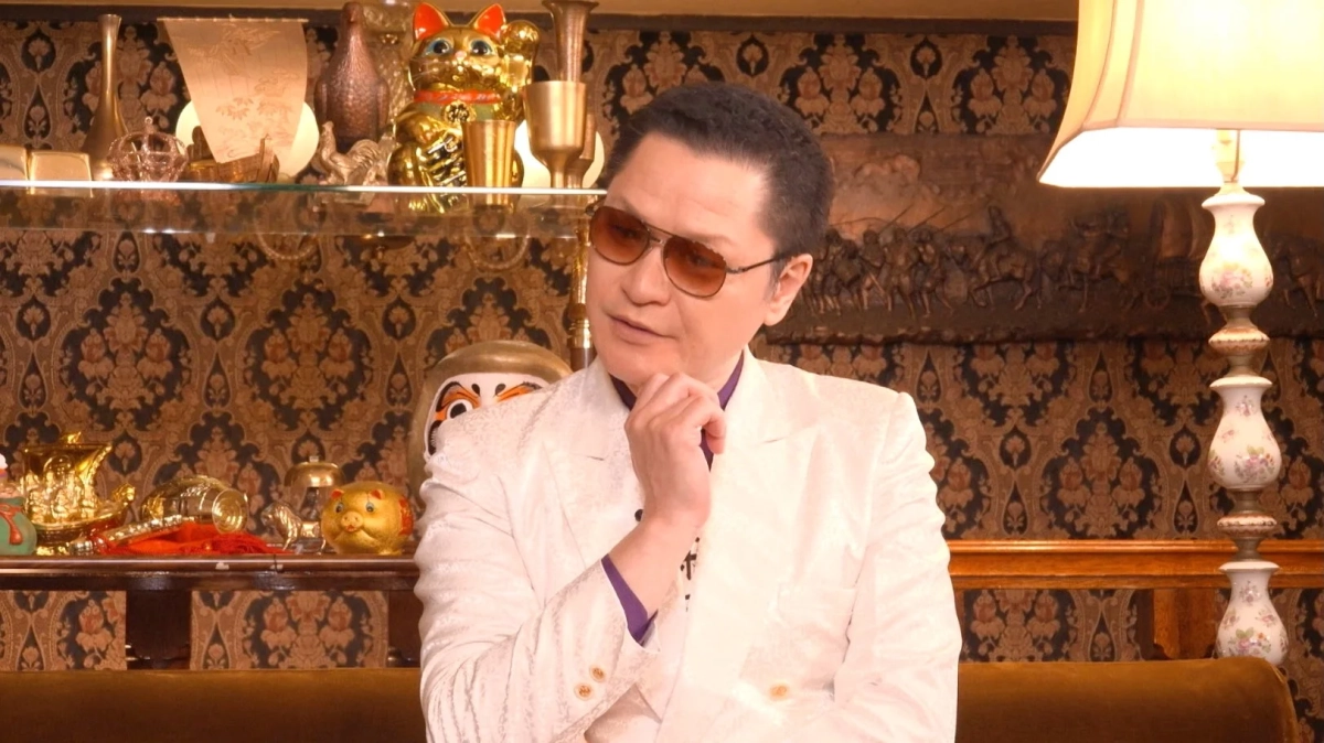 白いスーツとサングラスの男性が豪華な部屋で何かを考え込む様子