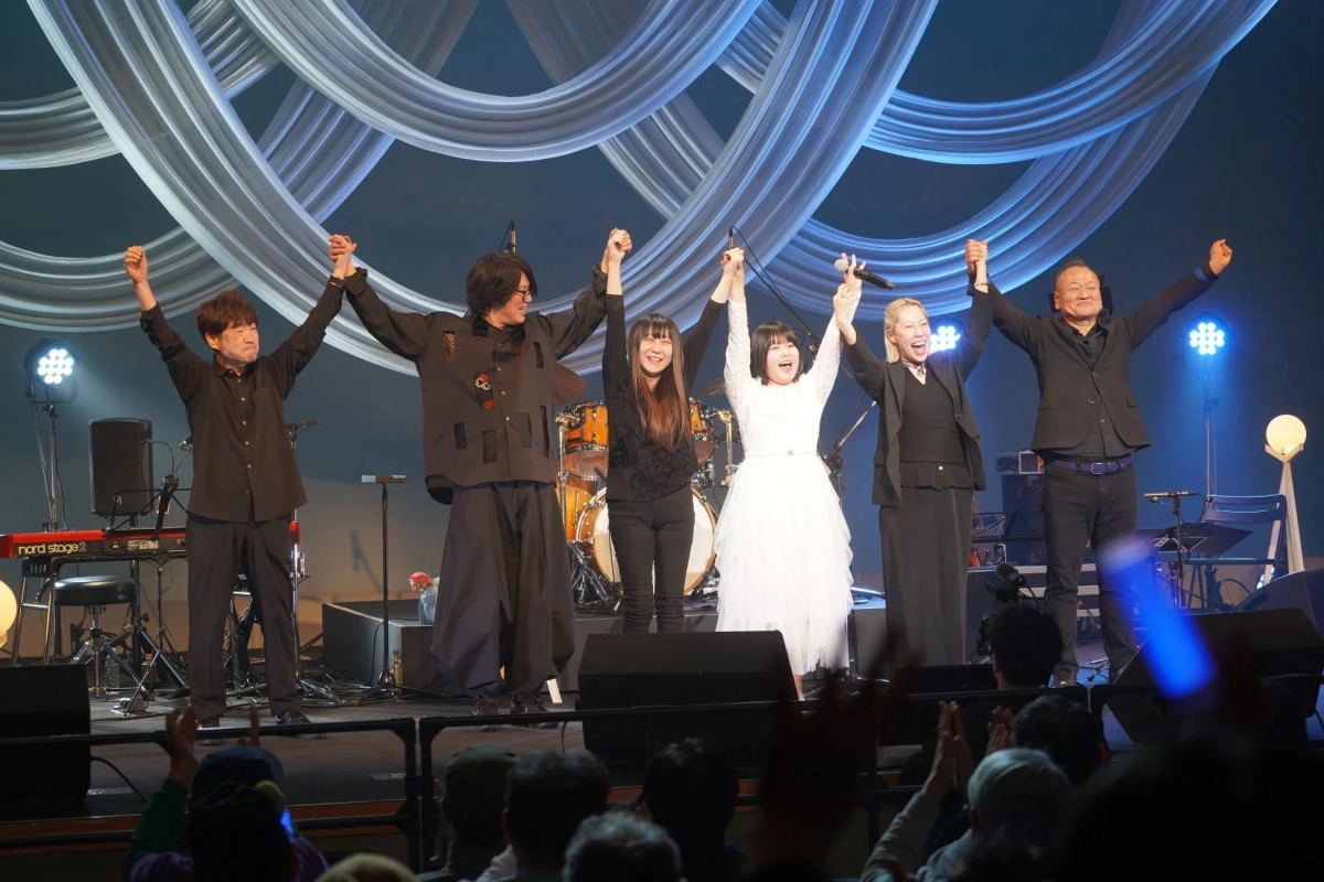 ライブステージで6人の出演者が手をつなぎ、手を上げて観客に挨拶しています