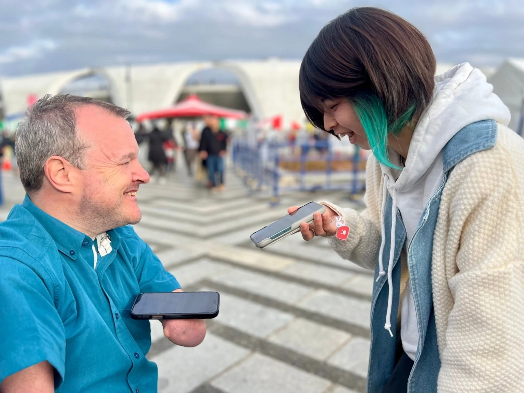 屋外で笑顔で会話する二人の人物。一人は腕に障がいを持つ男性、もう一人は緑のハイライトが入った髪の女性で、互いにスマートフォンを見せ合っている。