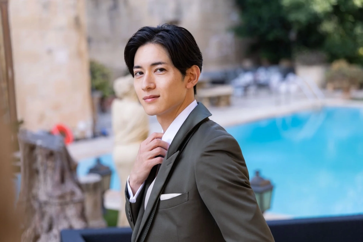 屋外のプールサイドで、若く端正な顔立ちの男性がカメラに向かって微笑んでいます。ダークグリーンのスーツを着ており、手で襟元を整える仕草をしています。洗練された雰囲気のポートレート写真です。