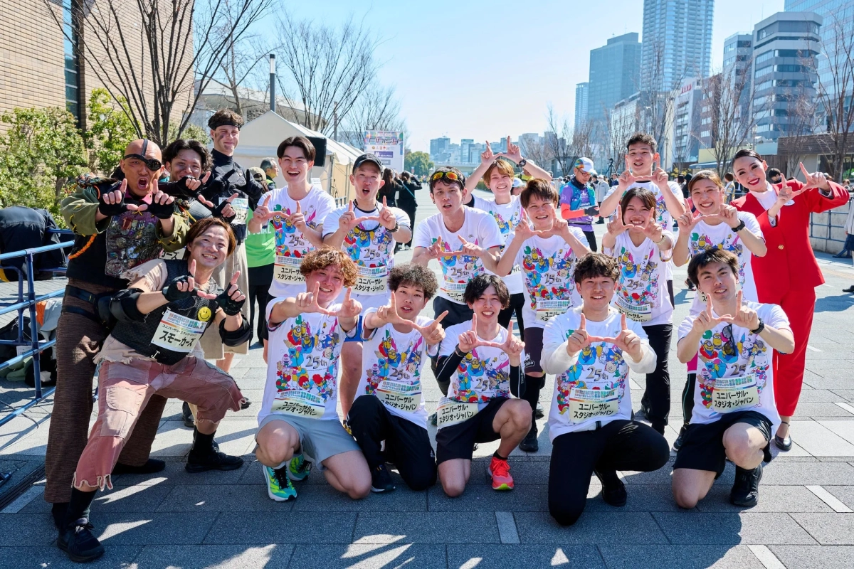 ユニバーサル・スタジオ・ジャパンの25周年を祝う屋外イベントで、参加者たちが笑顔でポーズを取っている集合写真です。キャラクターが描かれたTシャツを着ており、都市の晴れた日に開催されている様子が伺えます。