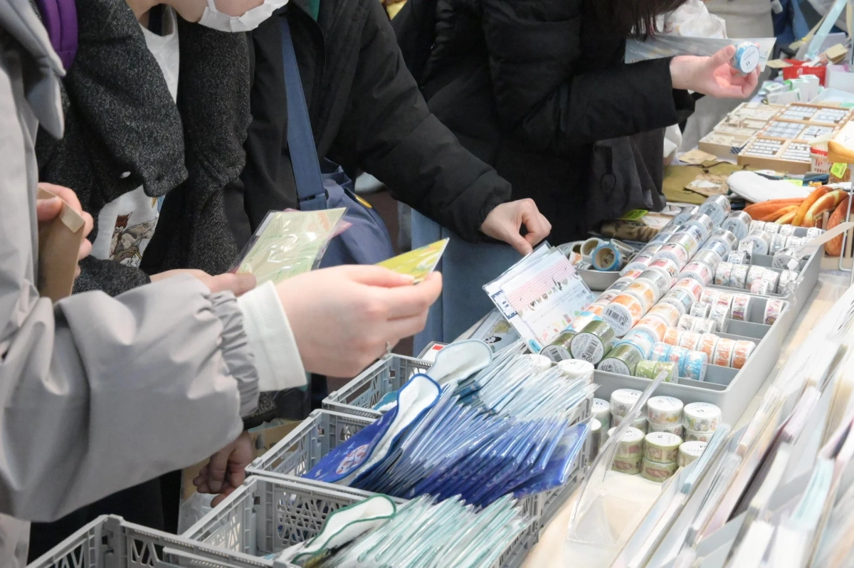 屋内のイベント会場か店舗で、人々がマスキングテープやその他の雑貨を選んでいる様子