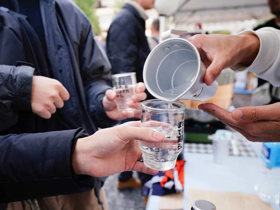 屋外イベントで、金属製のピッチャーから透明な液体がグラスに注がれている様子が写っています。