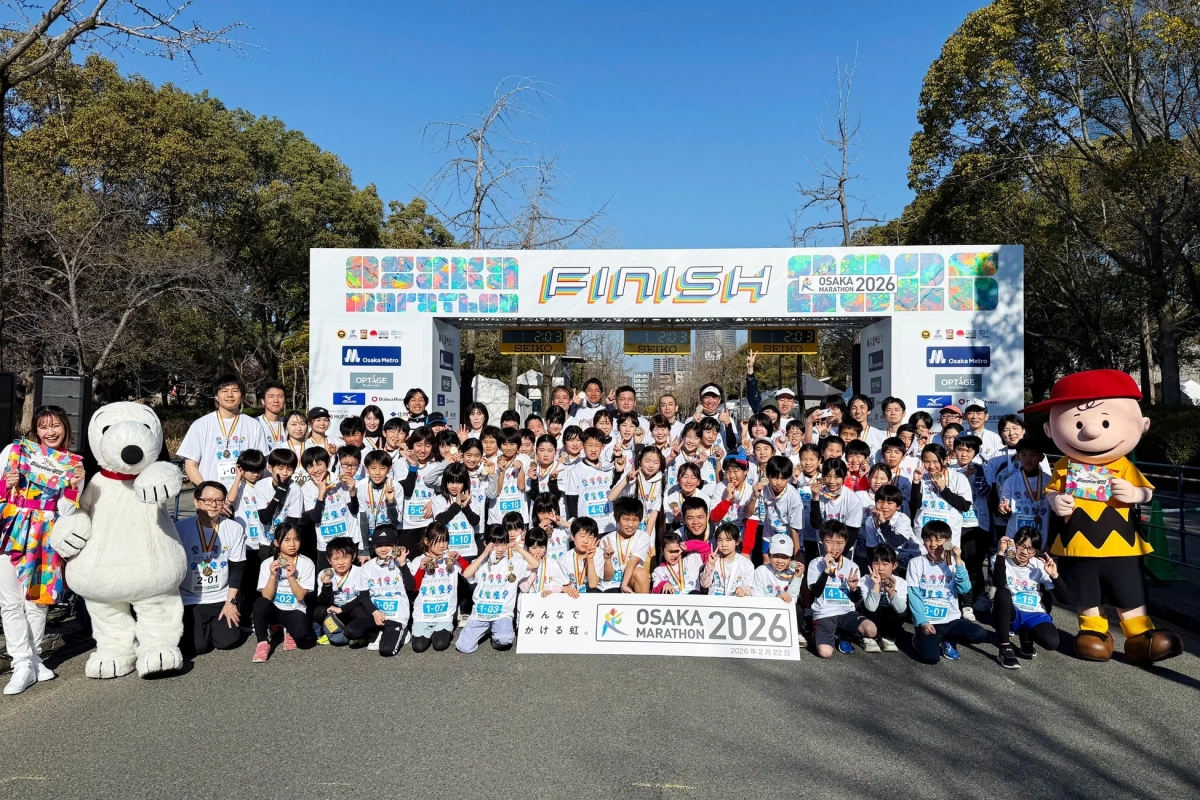 大阪マラソン2026のフィニッシュ地点で、大勢の子供たちが大人やスヌーピー、チャーリーブラウンのマスコットと一緒にメダルを手に笑顔で記念撮影をしている様子。