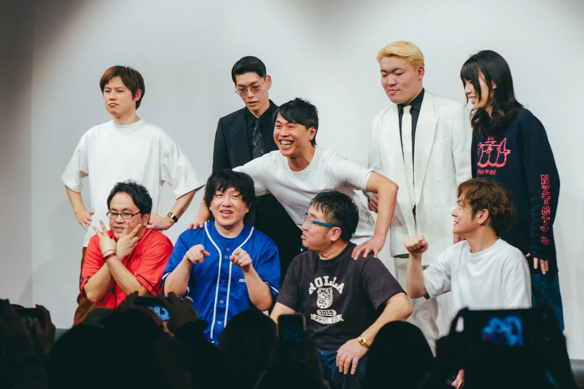 白い壁を背景に、男女9人が様々なポーズで並ぶ集合写真。笑顔やジェスチャーを見せる和やかな雰囲気