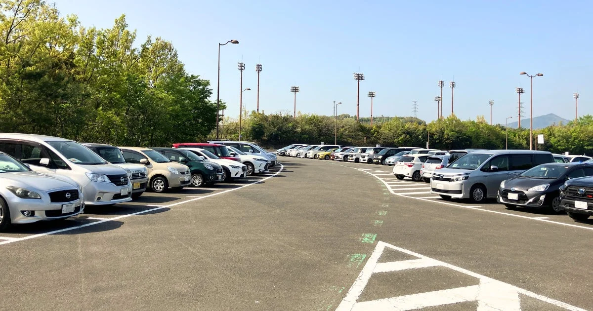 晴れた日に多くの車が整然と駐車している広々とした屋外駐車場です。背景には豊かな緑の木々や照明塔が見え、遠くには山々が広がっています。