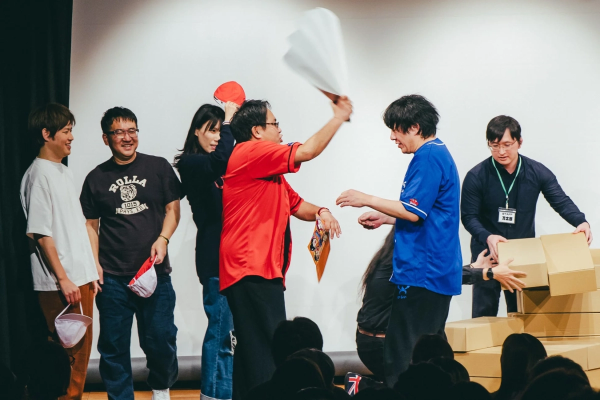 ステージ上で複数の人々が笑顔で交流しているイベント風景。赤いシャツの男性が白い紙を振り、青いシャツの男性と向き合っている。