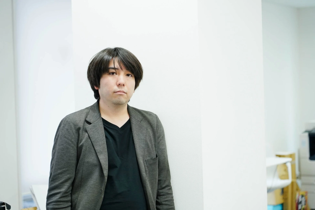 白い壁を背景に、グレーのジャケットと黒いVネックシャツを着用したアジア人男性が立っています。真剣な表情でカメラを見つめており、オフィスのような屋内環境が写し出されています。