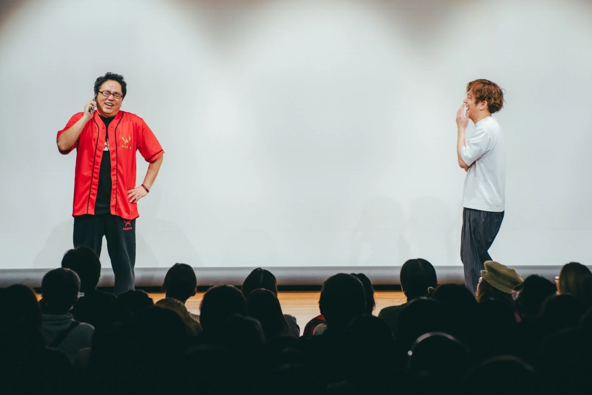 白い背景のステージ上で2人の男性が観客を前にパフォーマンスを行っています。赤いシャツの男性は電話を耳に当てて笑っており、もう一人の白いTシャツの男性も口元を抑えて笑っています。