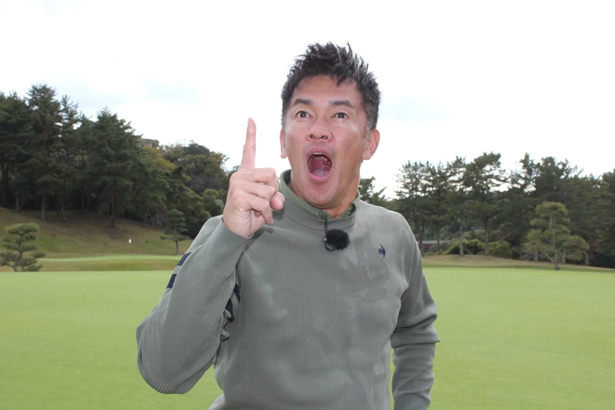 ゴルフ場に立つ男性が、口を大きく開けて驚いた表情で、人差し指を上に向けている様子を捉えた画像です。