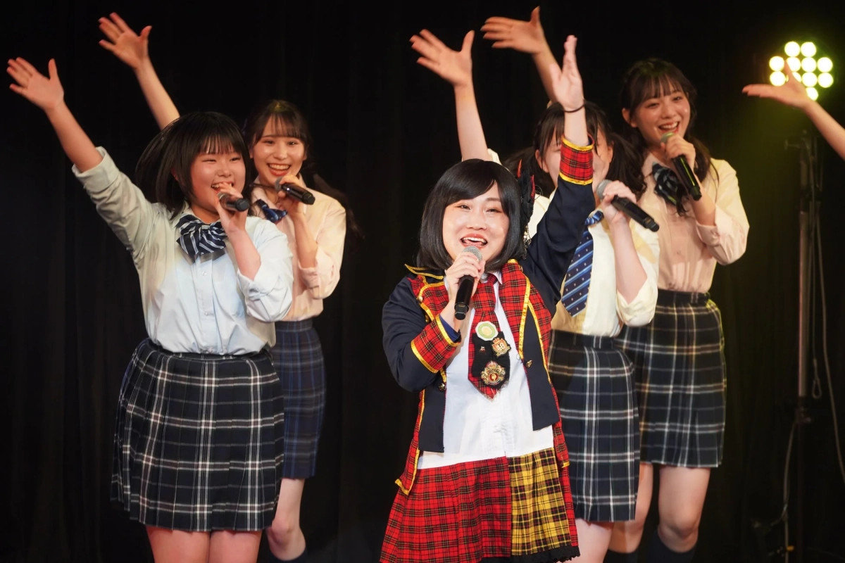 複数の若い女性がステージ上でマイクを持ち、笑顔で歌いながら手を上げているパフォーマンスの様子
