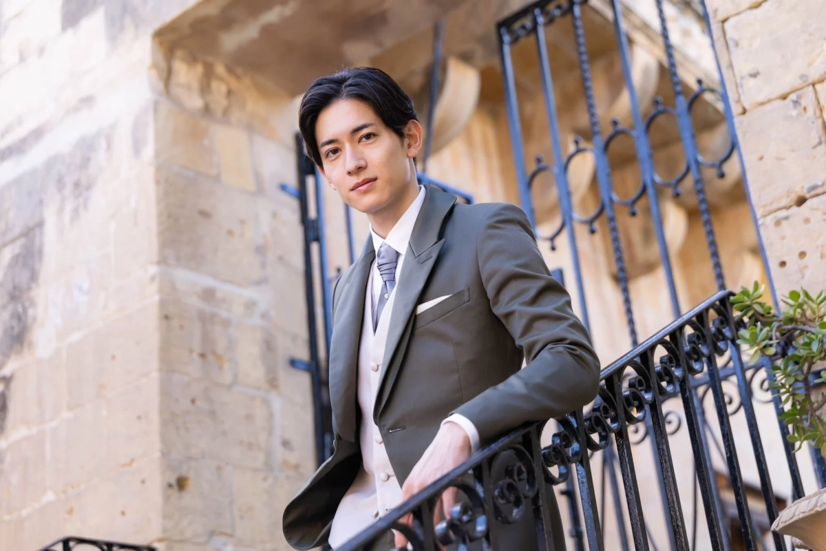オリーブグリーンのスーツを着用した若いアジア人男性が、歴史的な石造りの建物と装飾的な鉄柵を背景に立つポートレート。カメラを見つめる彼の落ち着いた表情が、上品で洗練された雰囲気を醸し出しています。