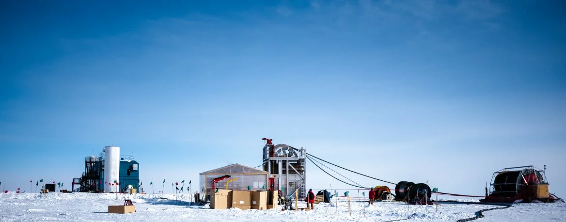 広大な雪原に立つ研究施設が広がる風景。青い空の下、複数の建物や観測機器、ケーブルが設置されており、数人の作業員が見られます。