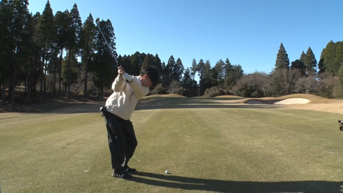 晴れた日のゴルフコースで、男性がゴルフのスイングをしている瞬間を捉えた画像です。緑豊かな芝生の上にはゴルフボールがあり、背景には木々やバンカーが見えます。青空が広がる開放的な雰囲気です。